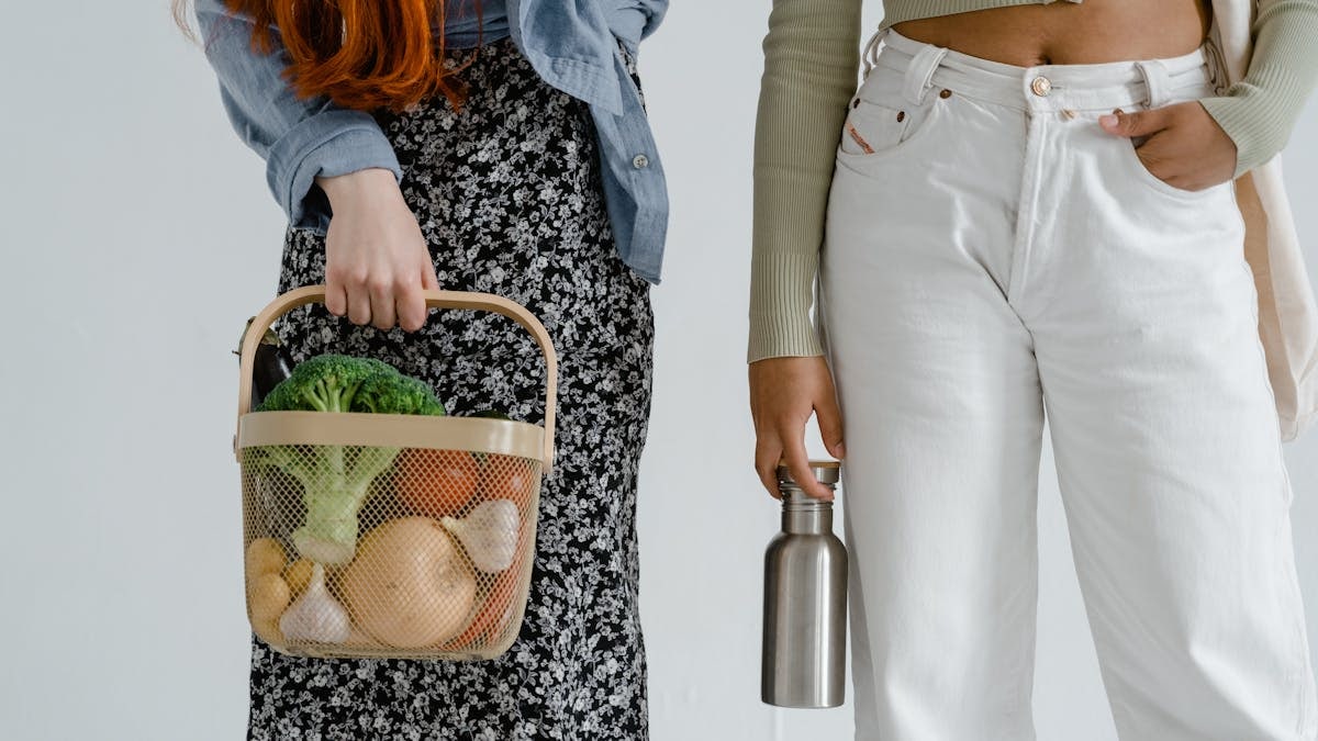 Two women holding a basket of fresh vegetables and a metal bottle, symbolizing sustainable lifestyle choices.